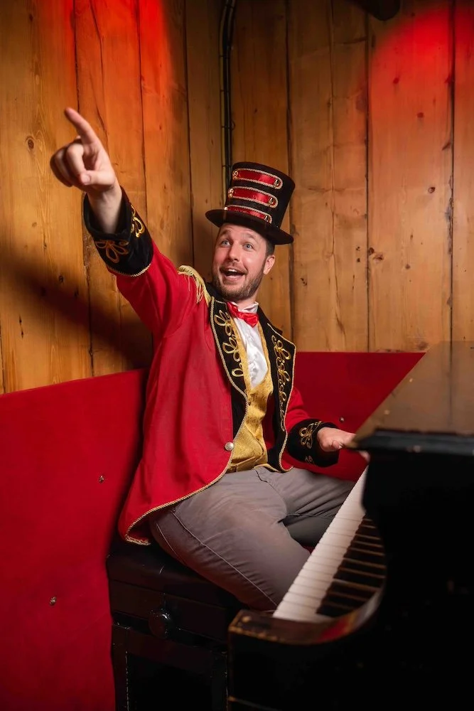 Performer in a red ringmaster costume at a piano, pointing out at the crowd