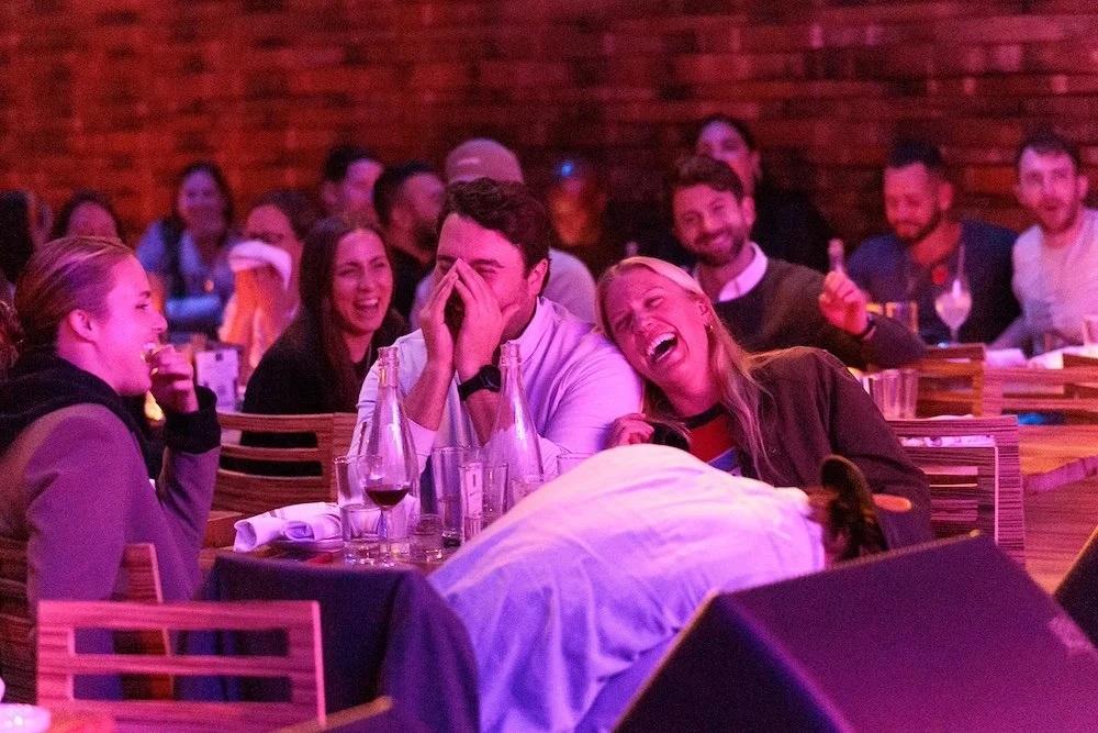 Audience howling with laughter at tables in a brick-walled comedy venue