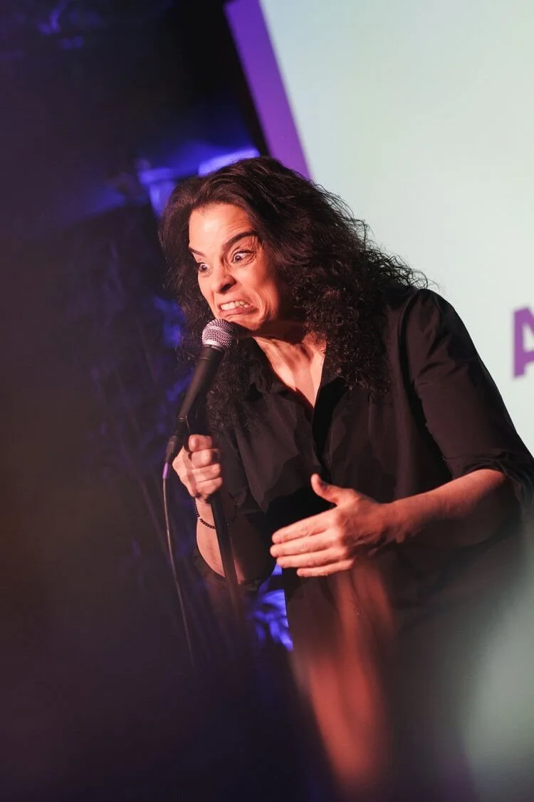 Female stand-up comedian leaning into a microphone on a purple-lit stage