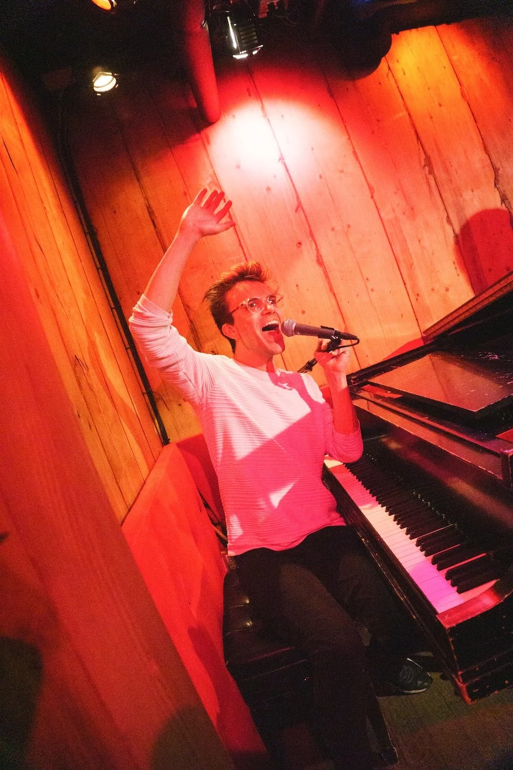 Musical comedian singing beside a grand piano under red stage lights