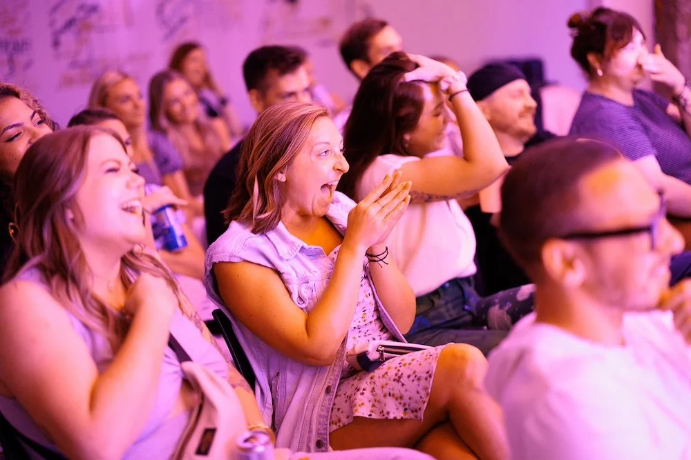 Rows of attendees reacting and laughing at a comedy show under purple lights