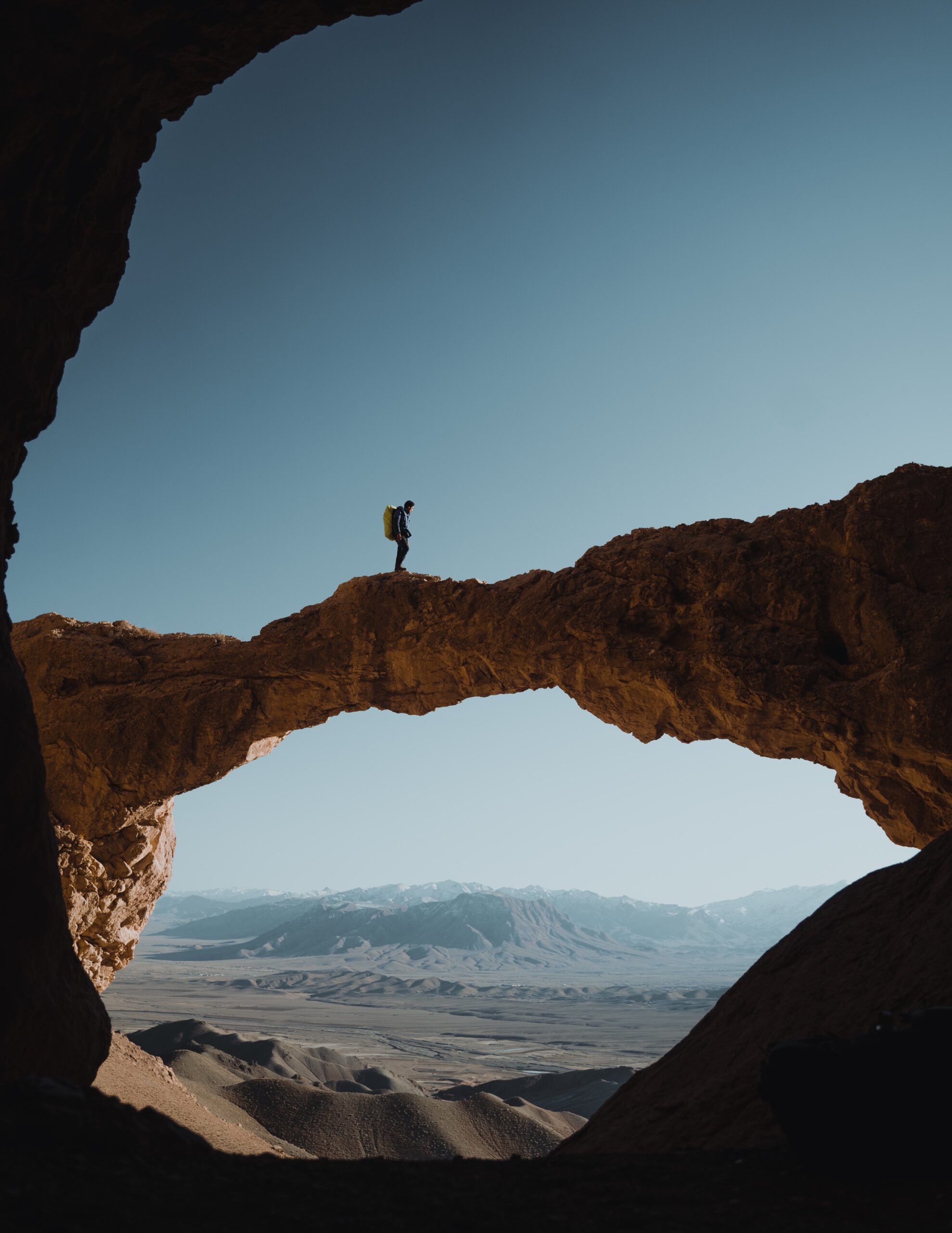 Hiker with a backpack standing on a natural rock arch above a vast desert valley with snow-capped mountains beyond