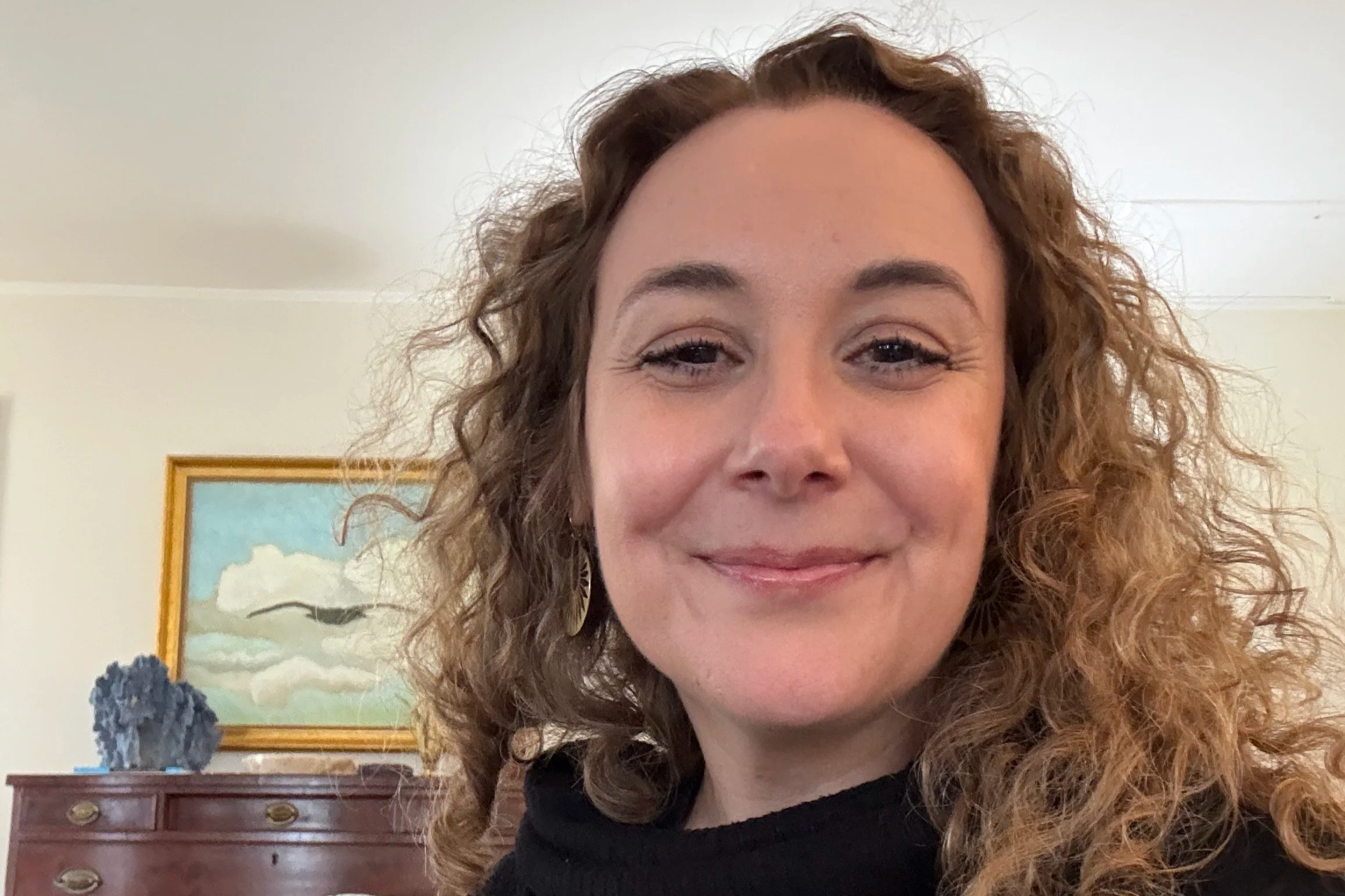 Nicole Bliss in a casual close-up, smiling indoors with a painting behind her