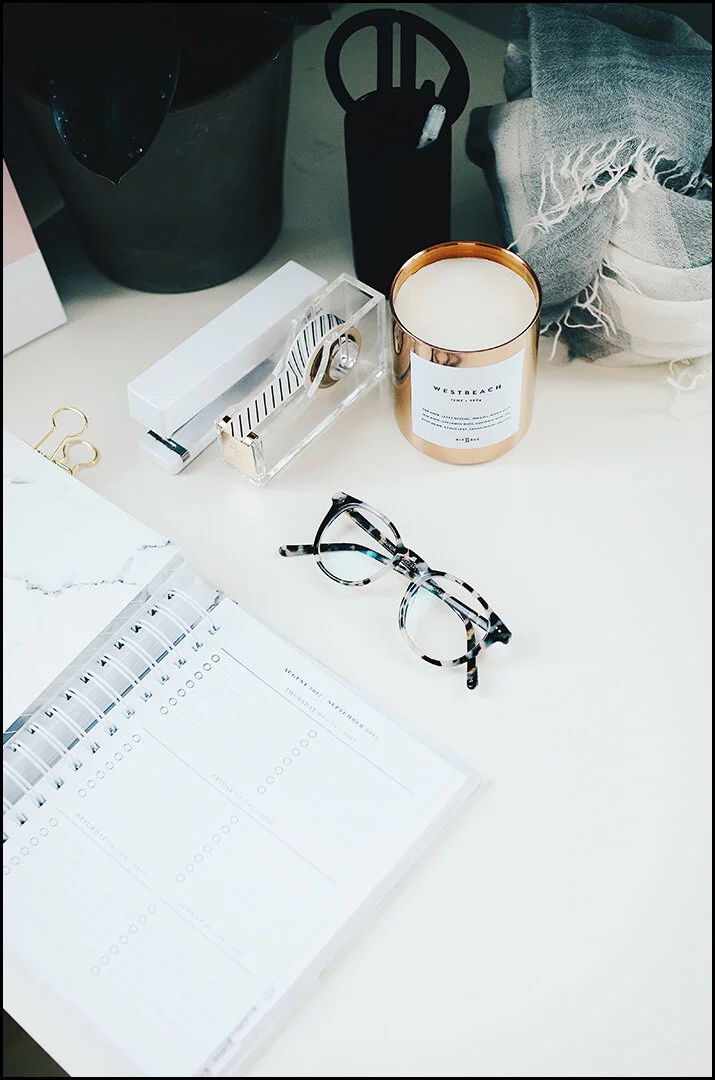 Styled desk flatlay with an open planner, glasses, candle and gold paper clips