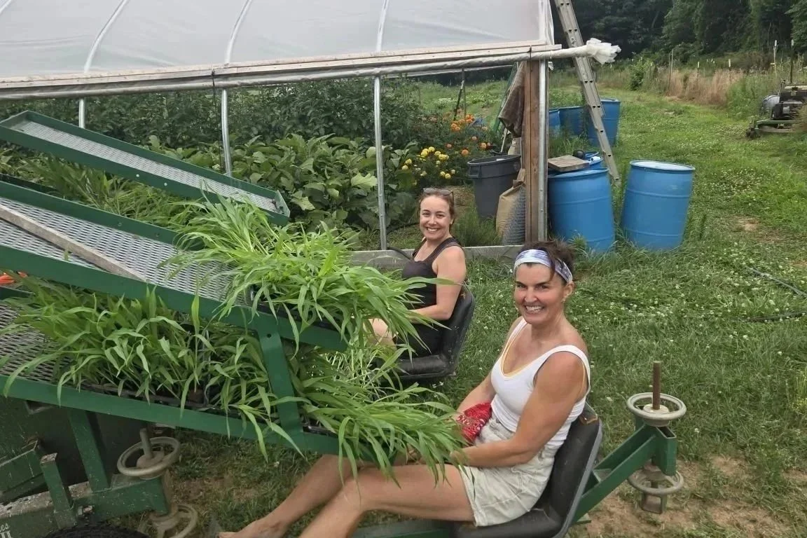 Two women smiling while seated on a farm transplanter beside a greenhouse
