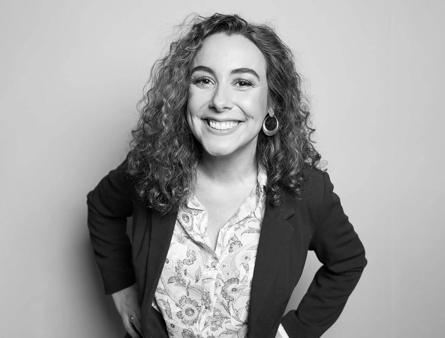 Nicole Bliss in a blazer and floral blouse, smiling with hands on hips — black and white portrait