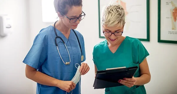 Two nurses in scrubs reviewing a tablet in a clinic hallway
