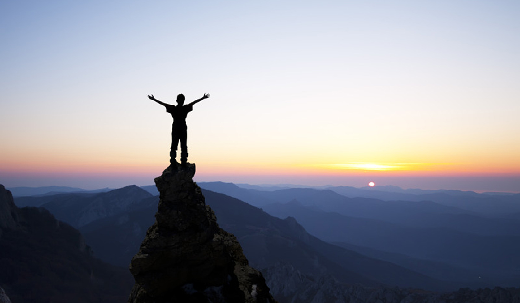 A person standing on a mountain summit with arms raised at sunrise