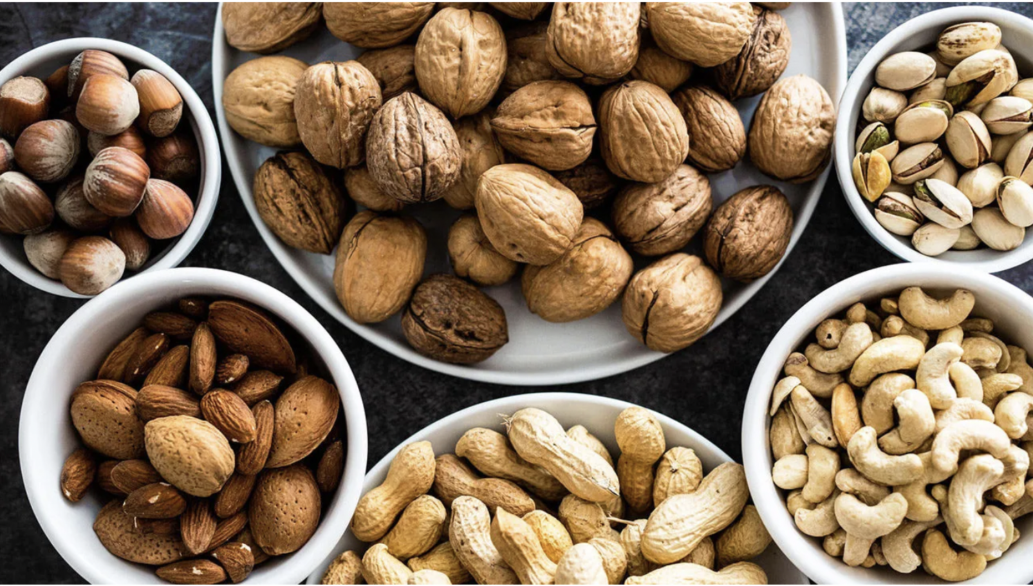Assorted nuts in white bowls, flat-lay on a dark surface