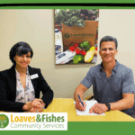 Two people signing a document at a table with the Loaves & Fishes logo
