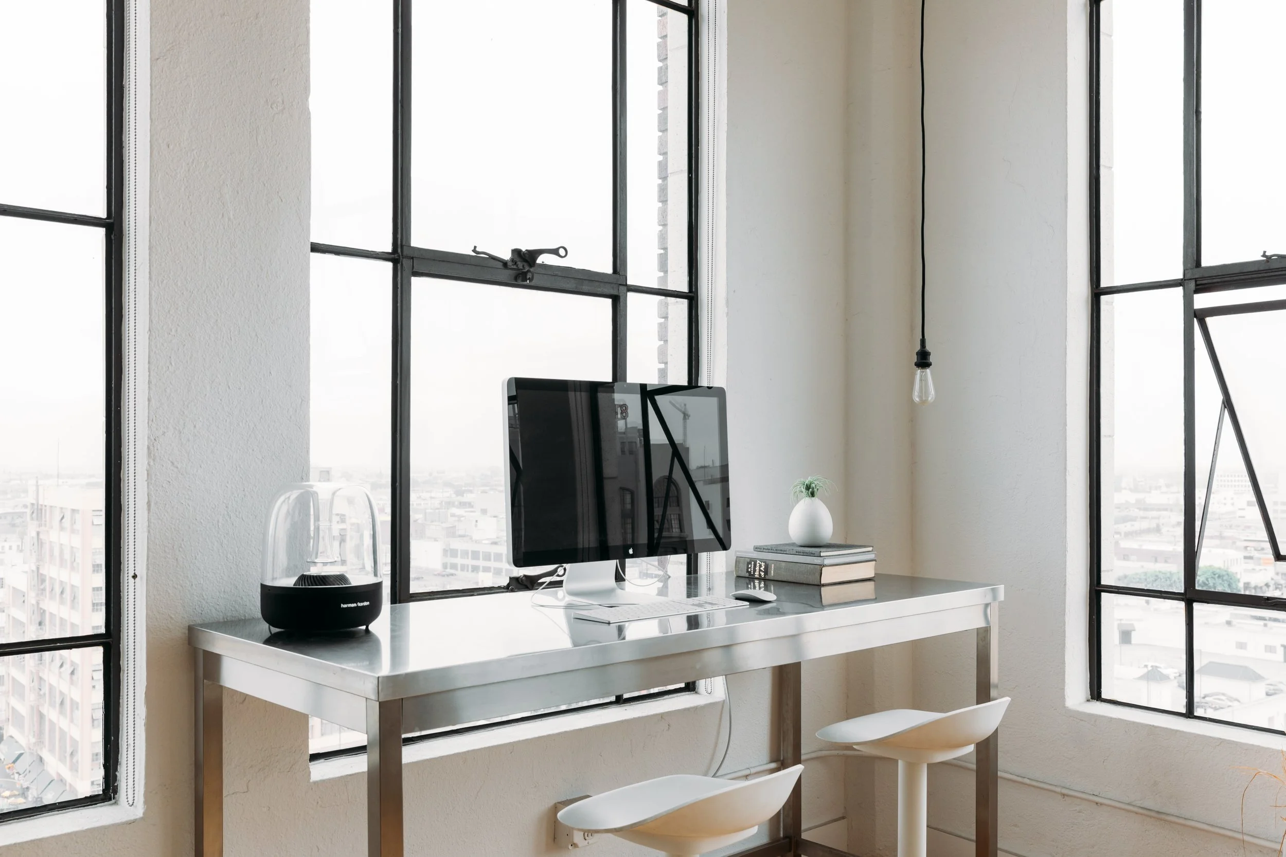 Loft workspace with an iMac on a metal desk beside large industrial windows overlooking the city