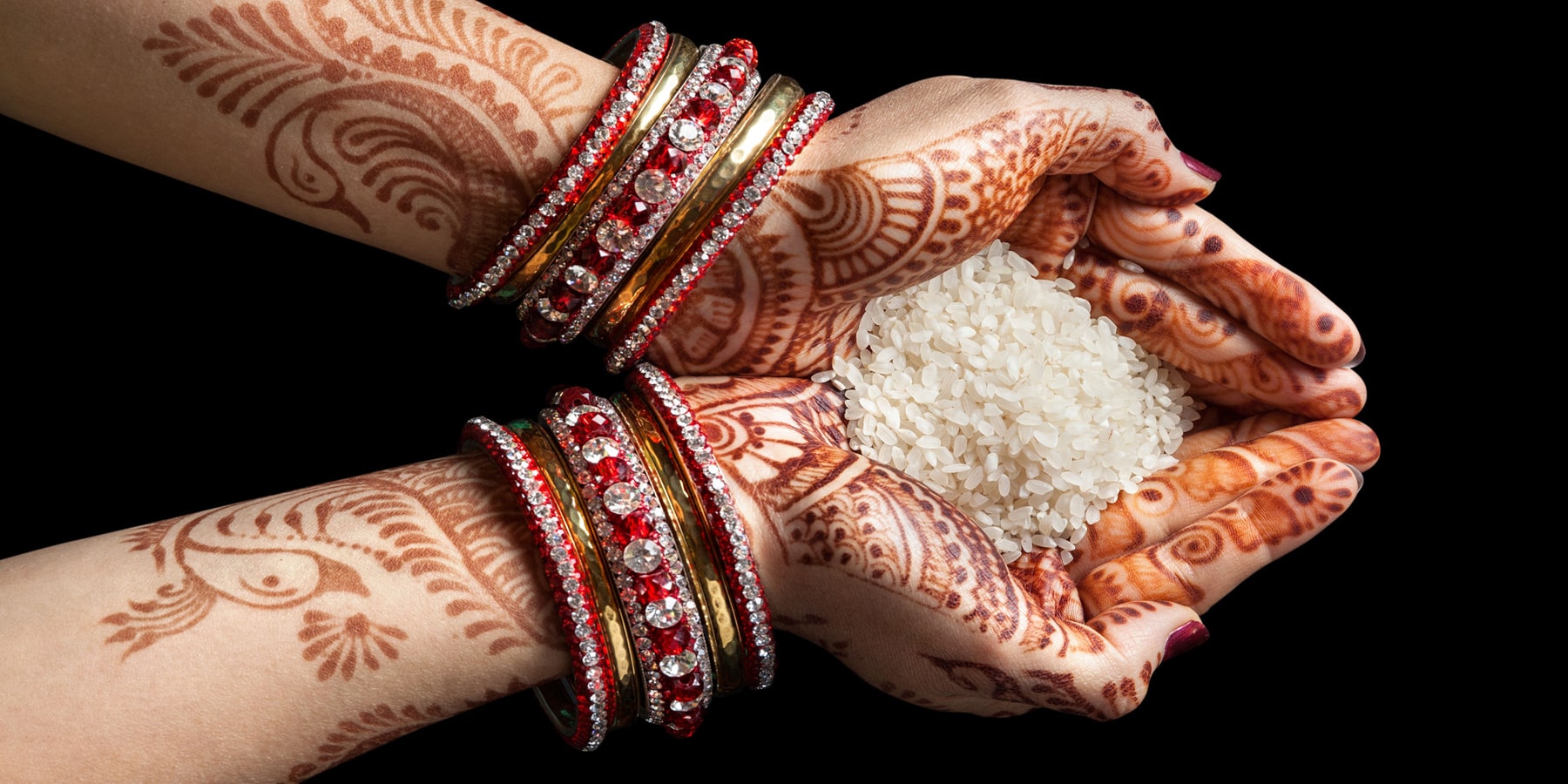 Close-up of two pairs of hands decorated with henna and red bangles, cupped together holding white rice