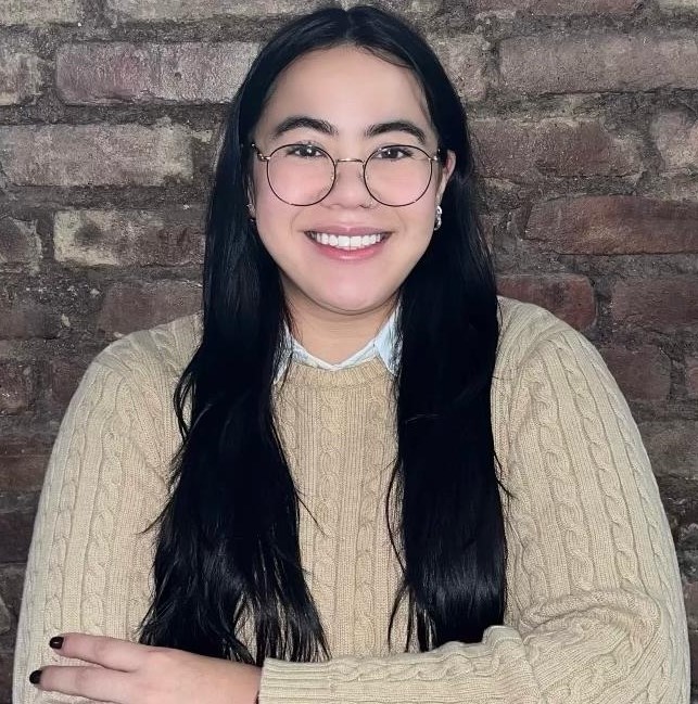 Portrait of Secia Umaña, smiling in a beige knit sweater against a brick wall