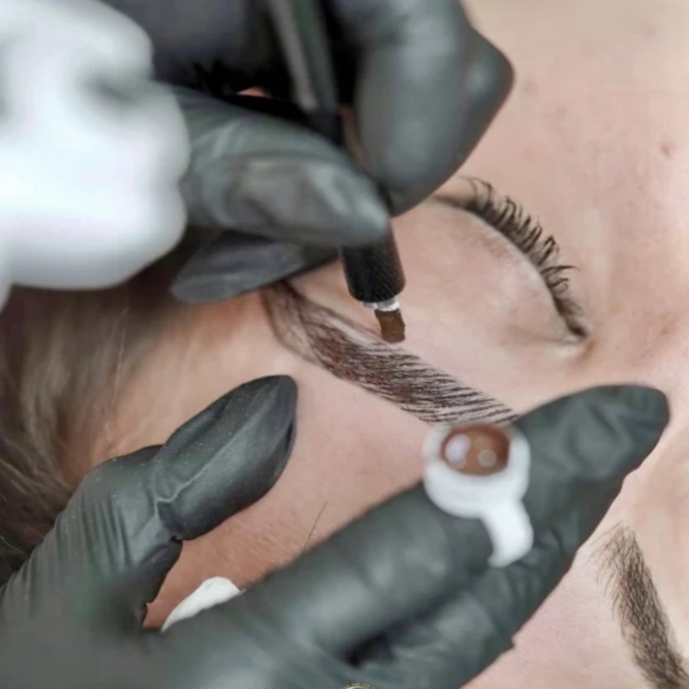Gloved hands performing nanoblading on a client's eyebrow