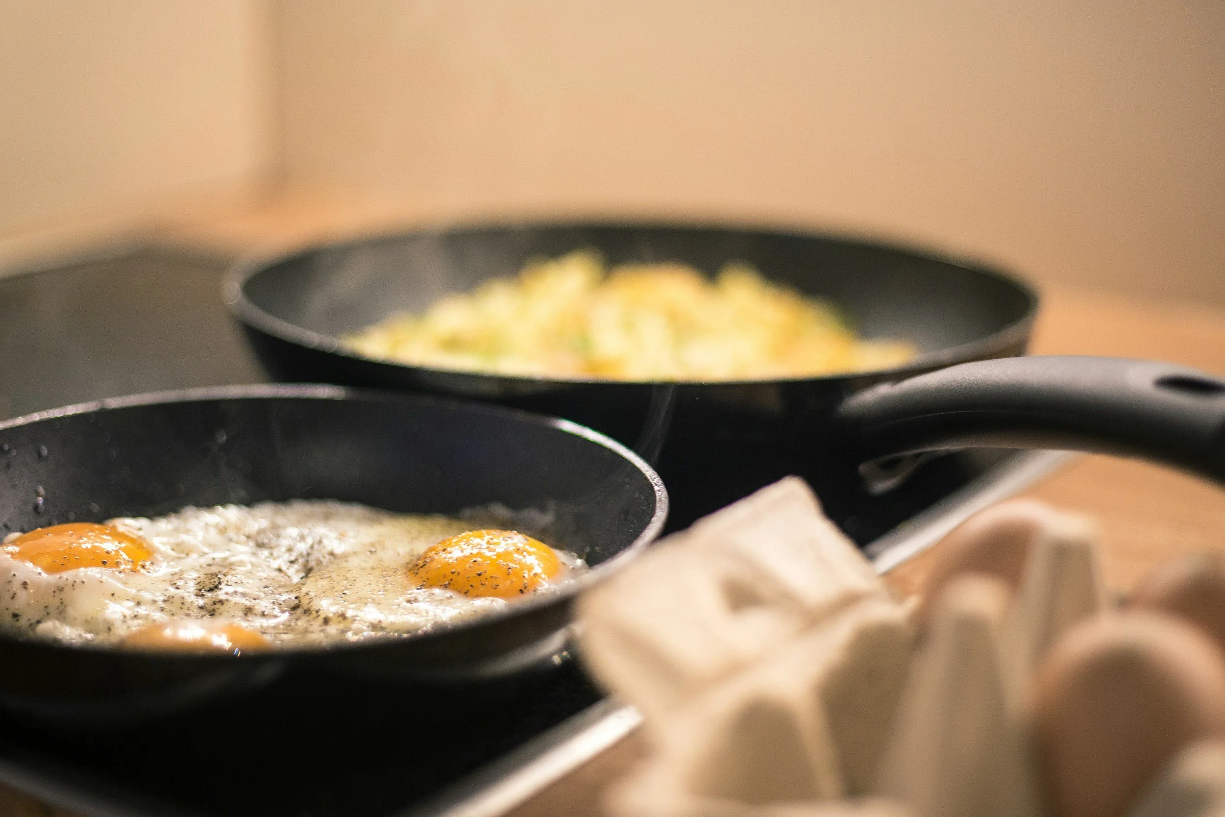 Two frying pans with eggs and potatoes cooking on a stovetop
