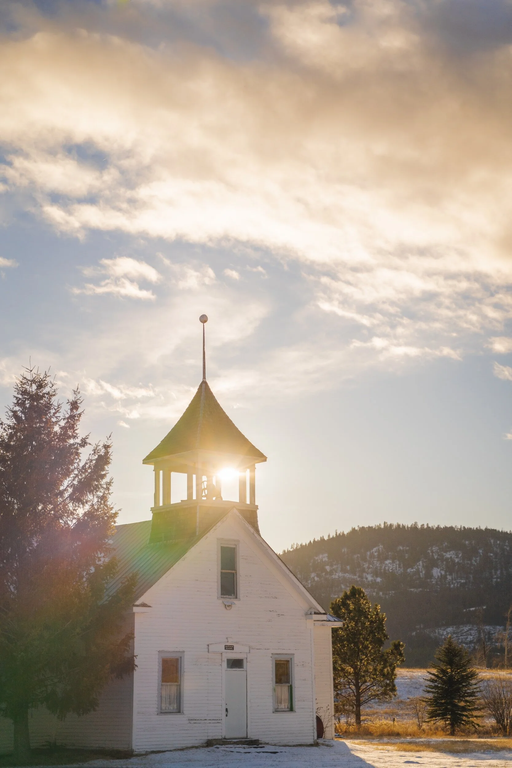 Small white country church with a bell-tower steeple, sun flare and snowy hills at golden hour