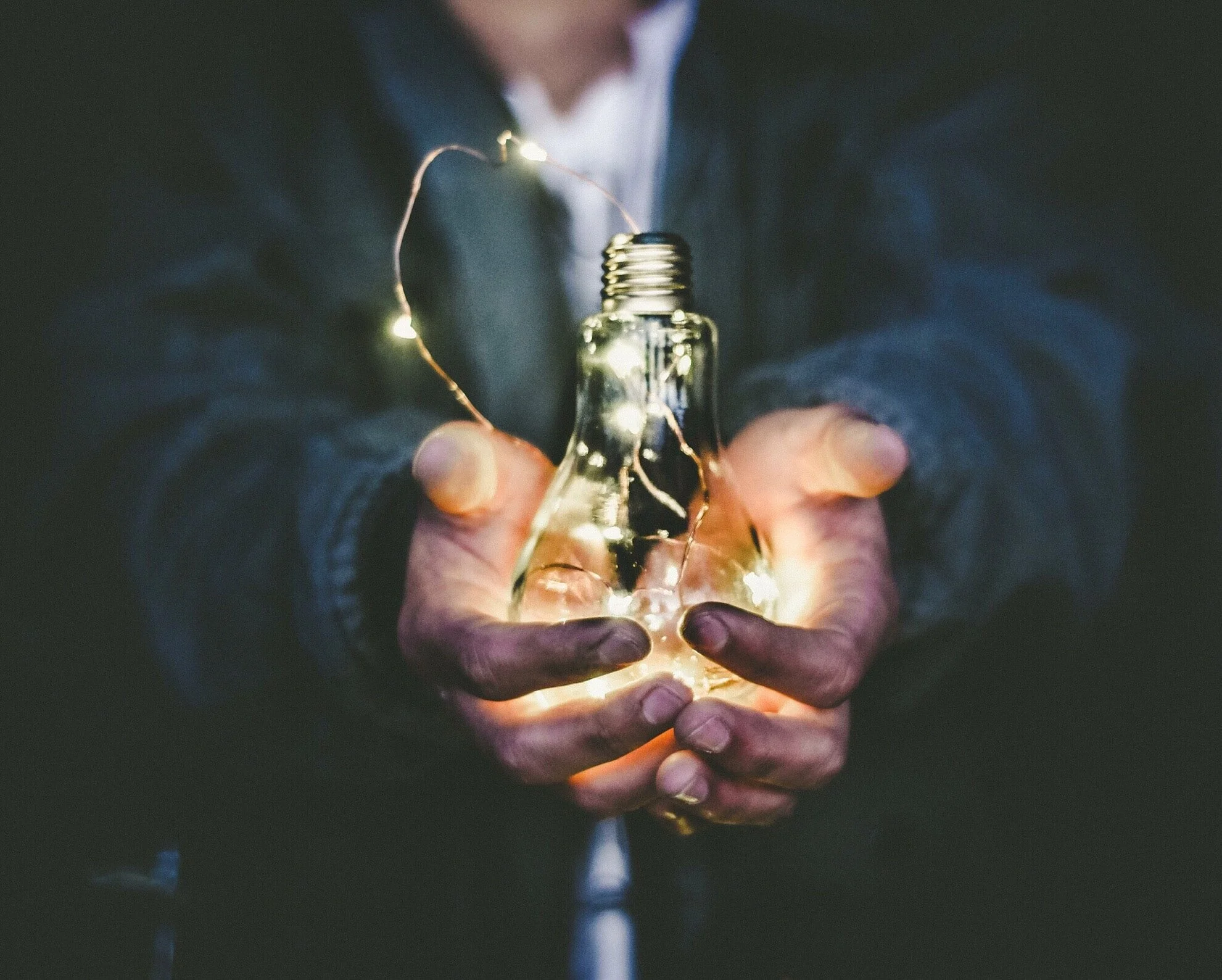Person in a suit holding a glowing light bulb wrapped in fairy lights in cupped hands
