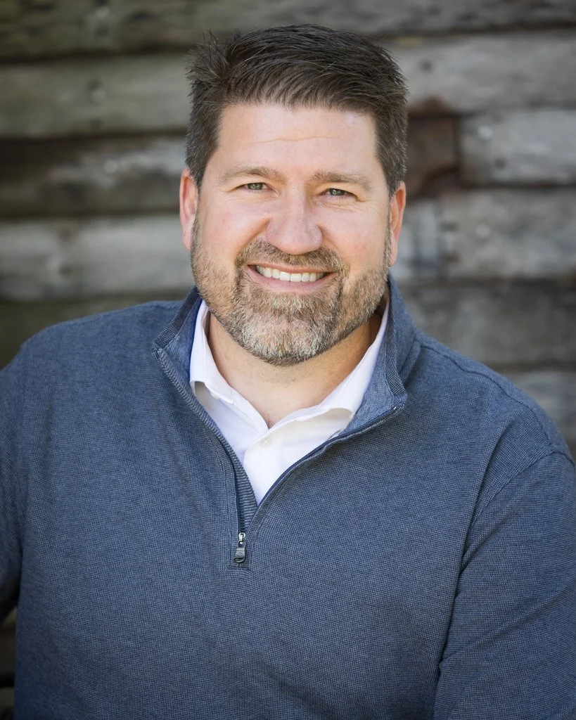 Heath Luehmann, a smiling middle-aged man with dark hair and a beard, wearing a blue quarter-zip over a white shirt