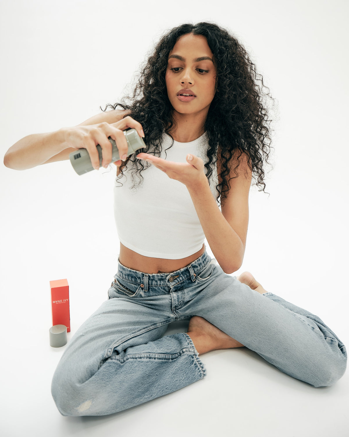 A woman with curly hair applying a Mane Ivy hair product, seated on a white floor.