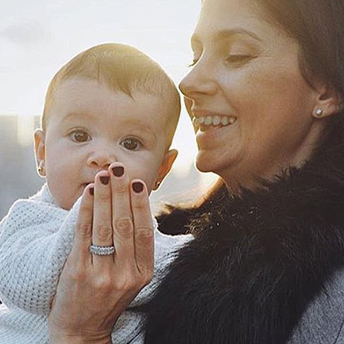 Smiling woman holding a baby, showing a dark burgundy manicure and diamond ring
