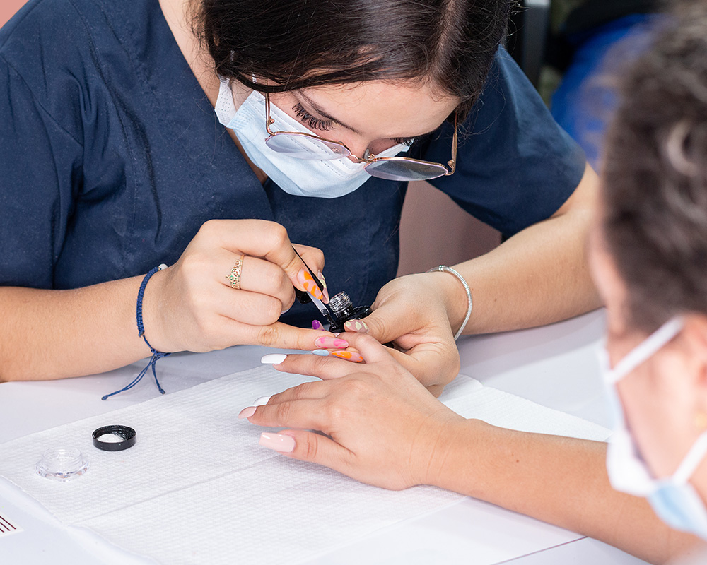 Masked nail technician in navy scrubs carefully painting intricate nail art
