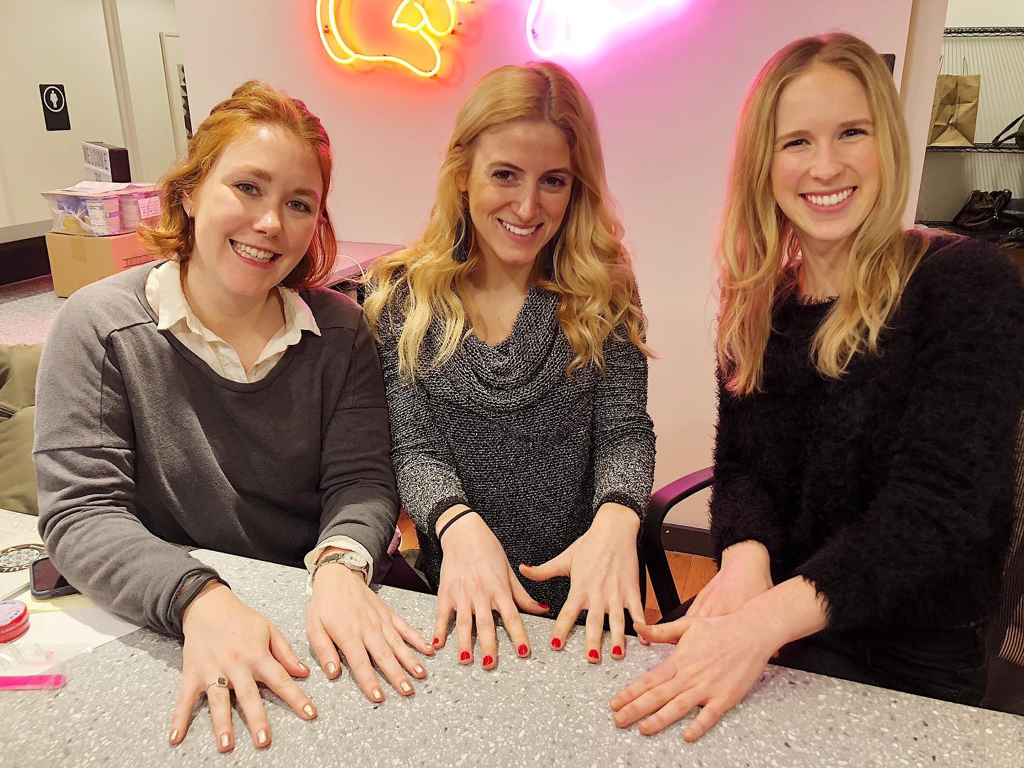 Three smiling women showing off freshly painted nails at a counter with a neon sign behind them