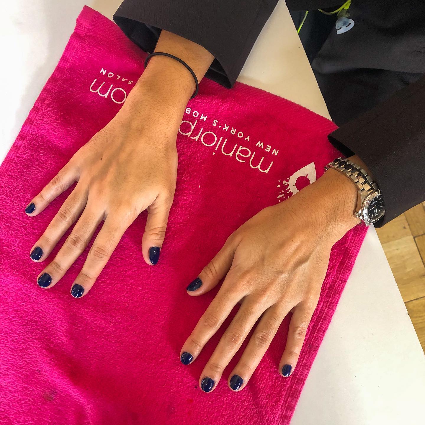 Two hands with dark navy manicure resting on a bright pink MANIORPEDI branded towel