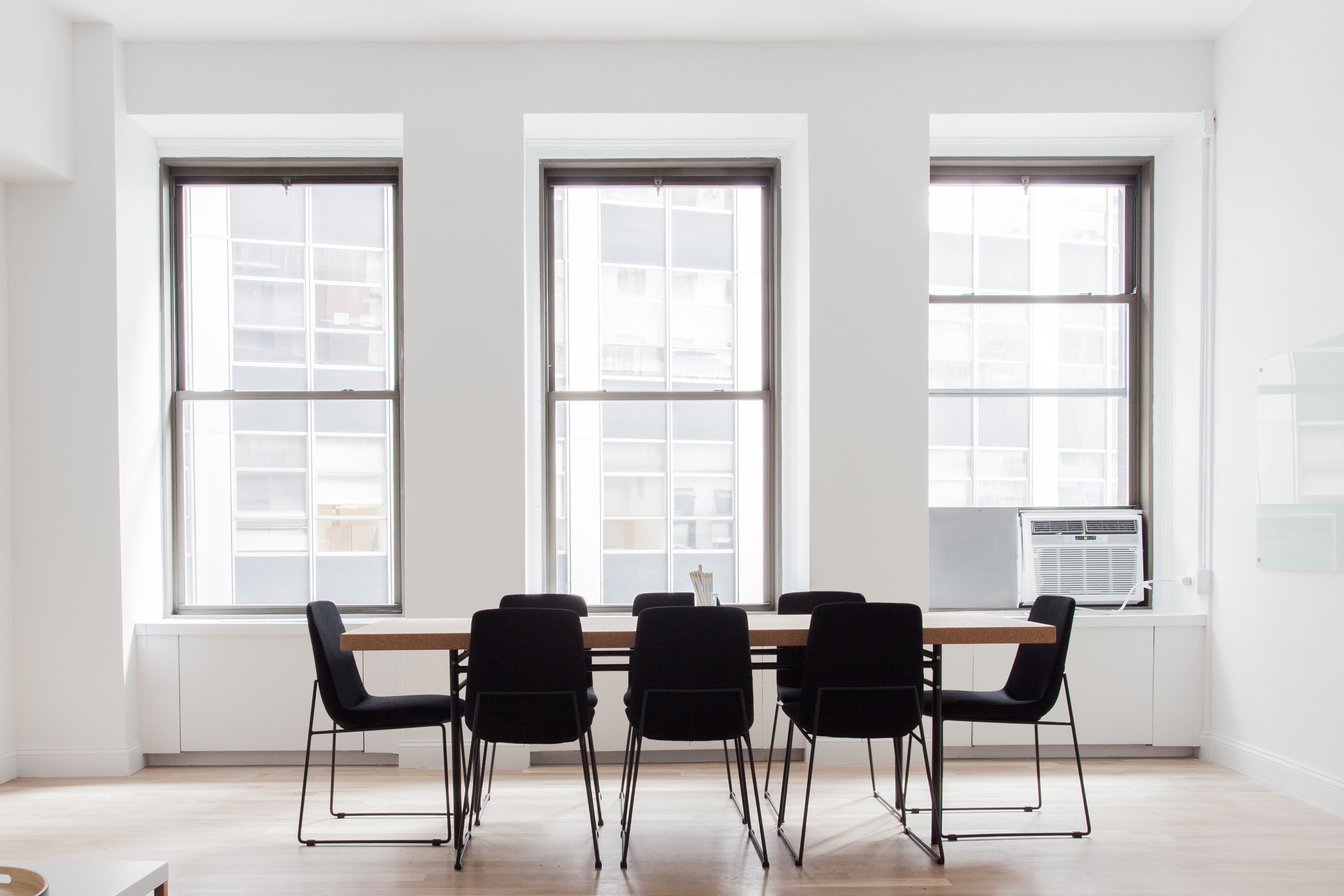 A minimalist conference room with a long wooden table, black chairs and three tall windows