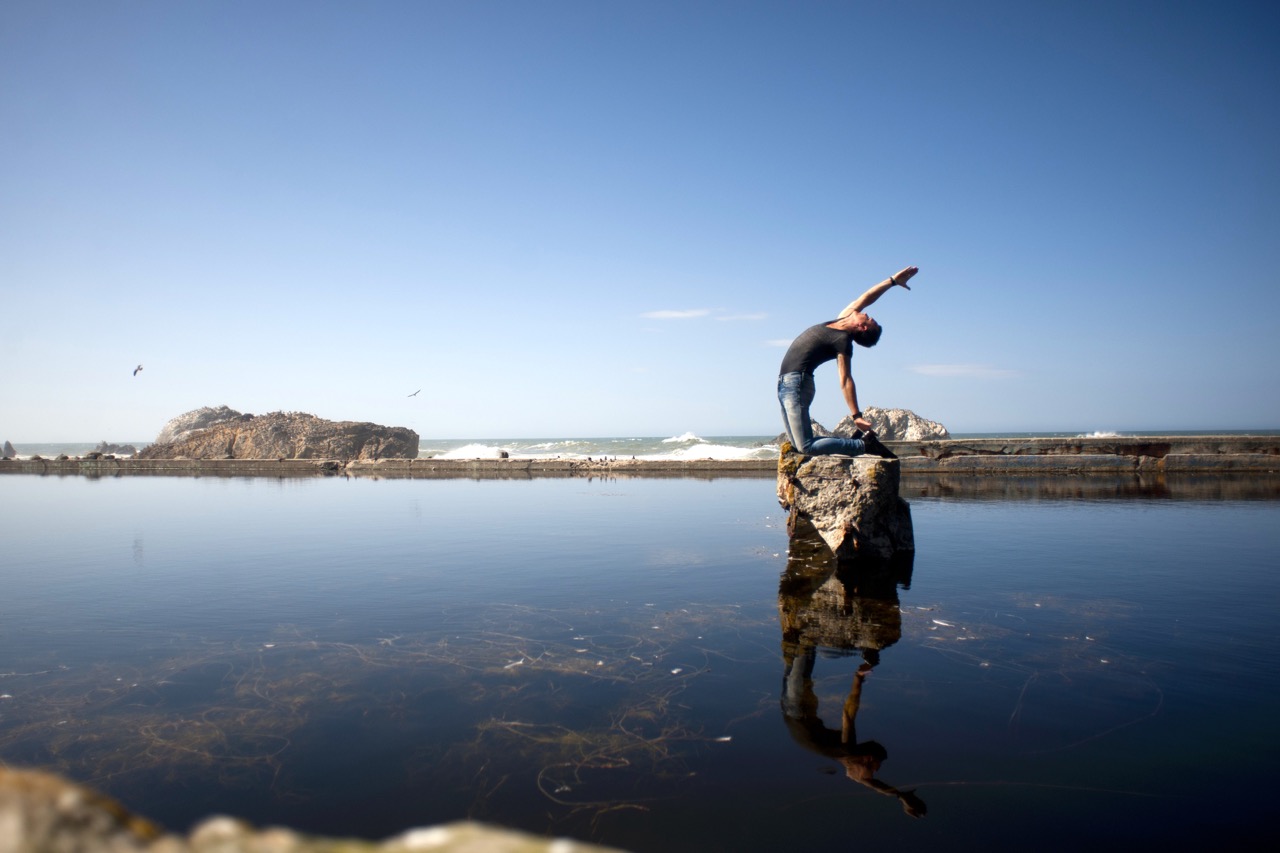 Mark Morford in a deep half-camel backbend on a rock at Sutro Baths, reflected in still water