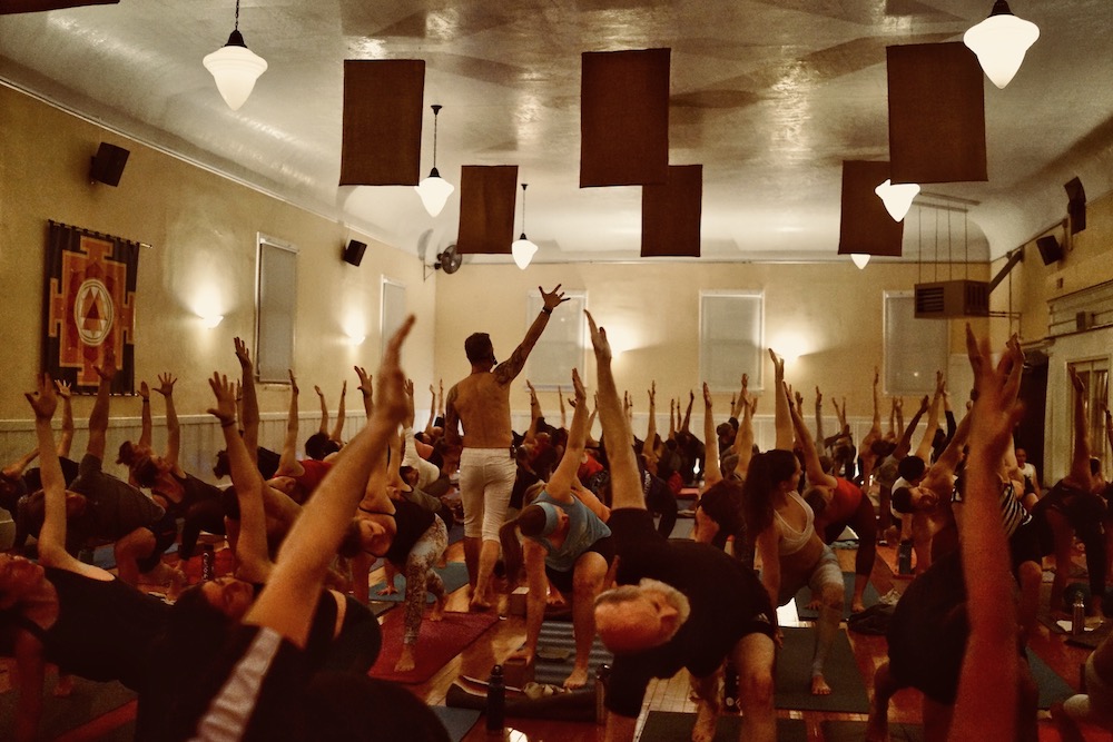 Large candlelit yoga class with instructor's arms raised at the front