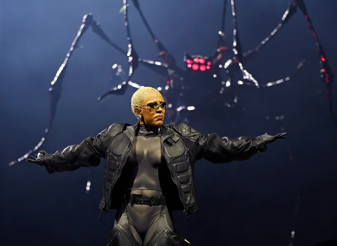 Performer with arms outstretched on a dark stage before a large spider prop backdrop