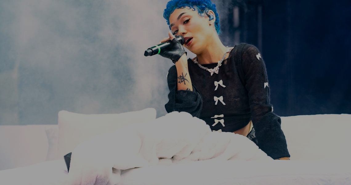 Artist with blue hair performing with a microphone on stage amid smoke