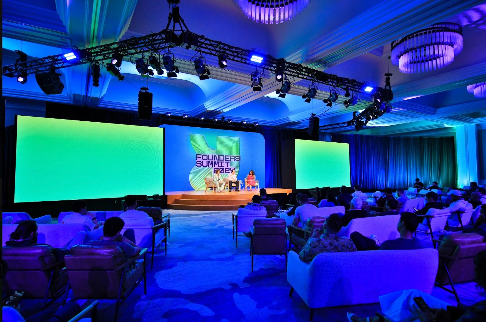 Indoor conference ballroom in blue light with a Founders Summit 2024 stage, large LED screens and a seated audience