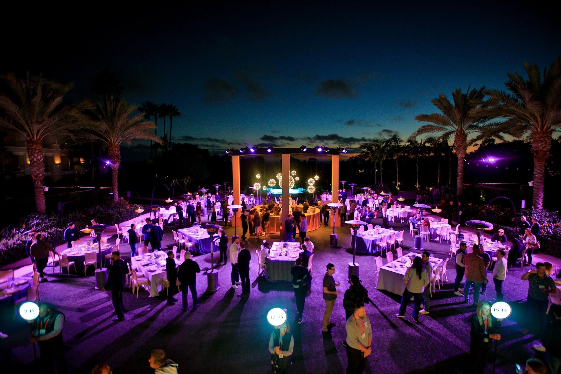 Outdoor corporate event at dusk with purple lighting, palm trees, round tables and hanging globe lights as guests mingle