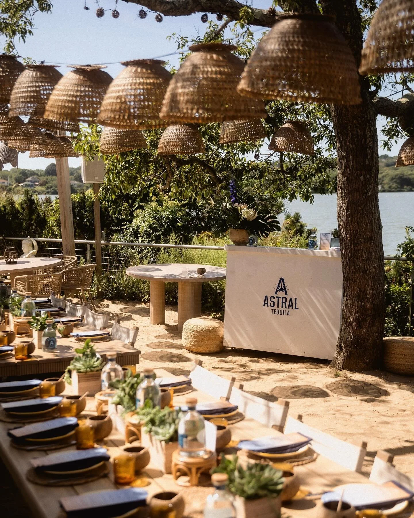 Outdoor Astral Tequila dinner setup under wicker pendant lights with a set table and waterside Hamptons backdrop