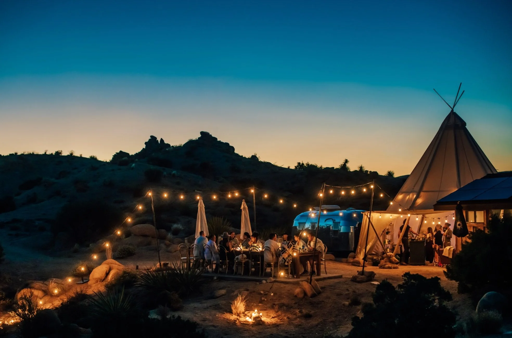 Twilight desert event with string lights, tipi tent, Airstream trailer and guests dining at a long table