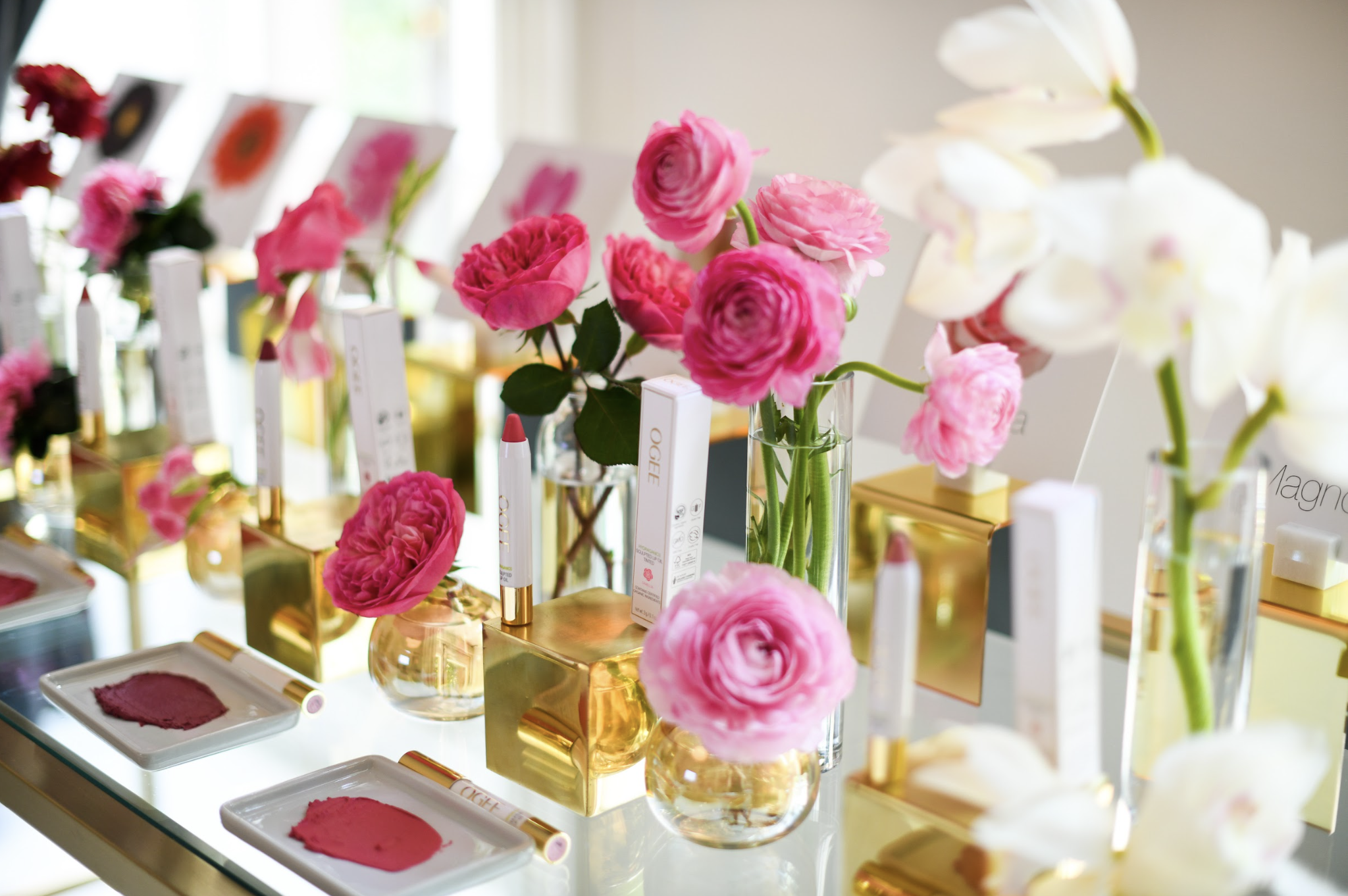 Beauty brand event display with pink roses and gold-accented Ogee lipstick products on glass shelving