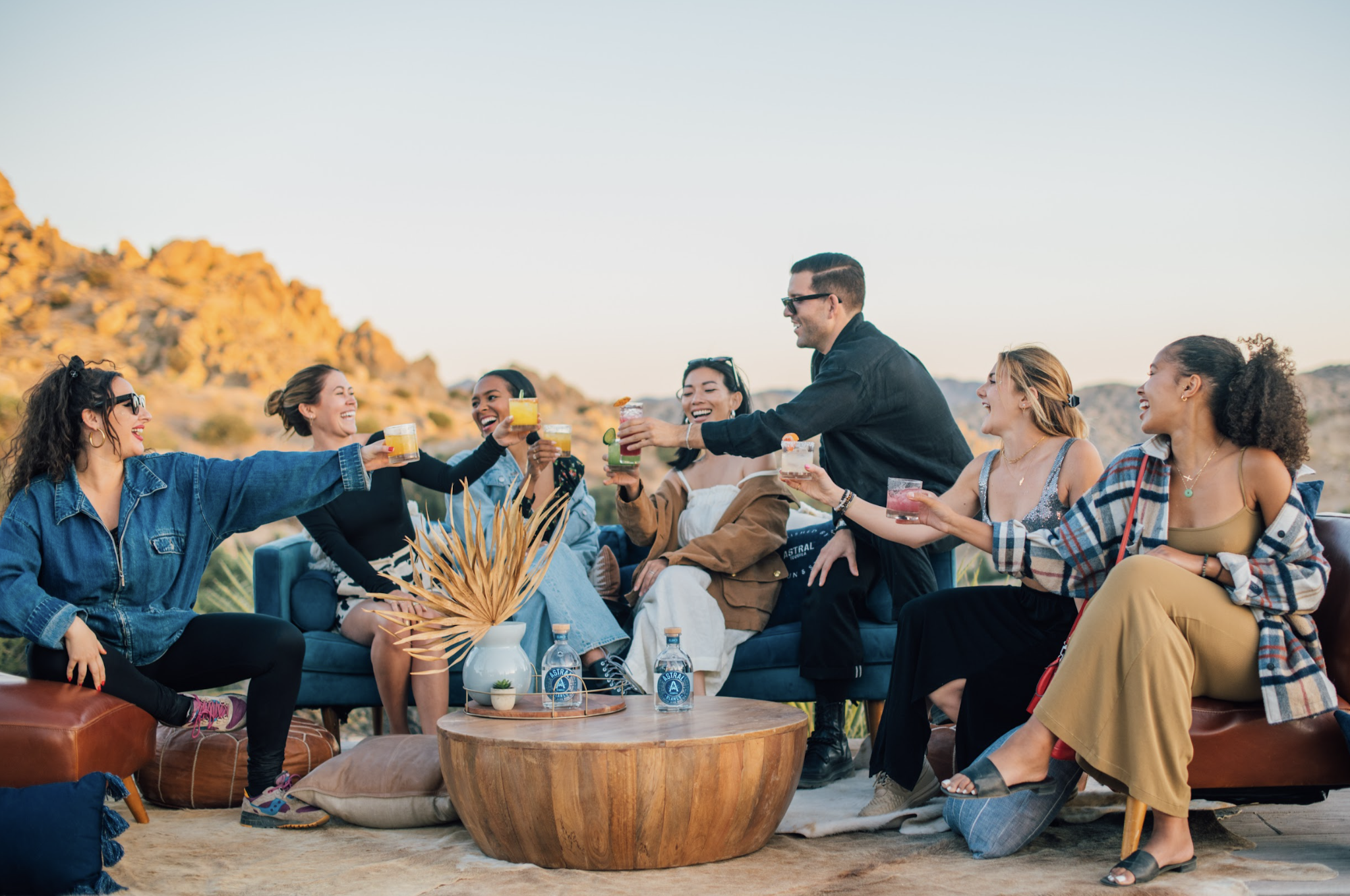 Group of seven people toasting cocktails outdoors in the Joshua Tree desert with Astral Tequila bottles on the coffee table