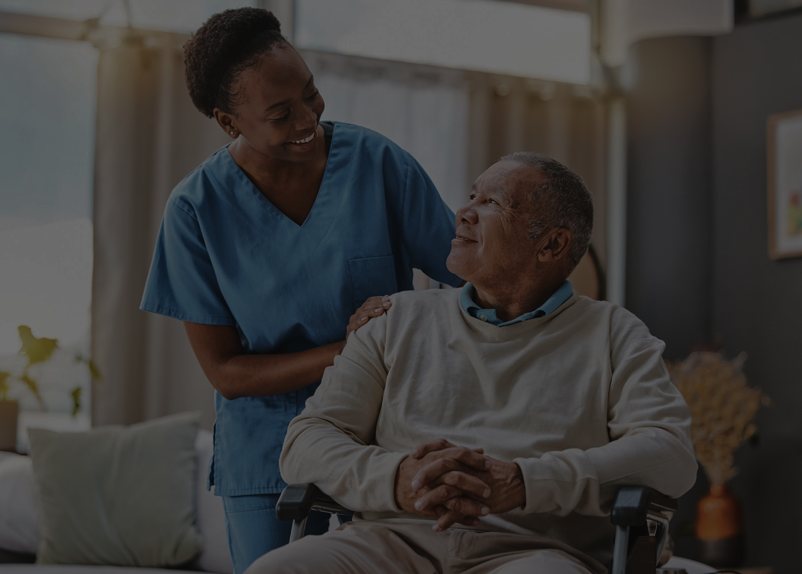 A nurse in blue scrubs smiling with an elderly man seated in a wheelchair at home