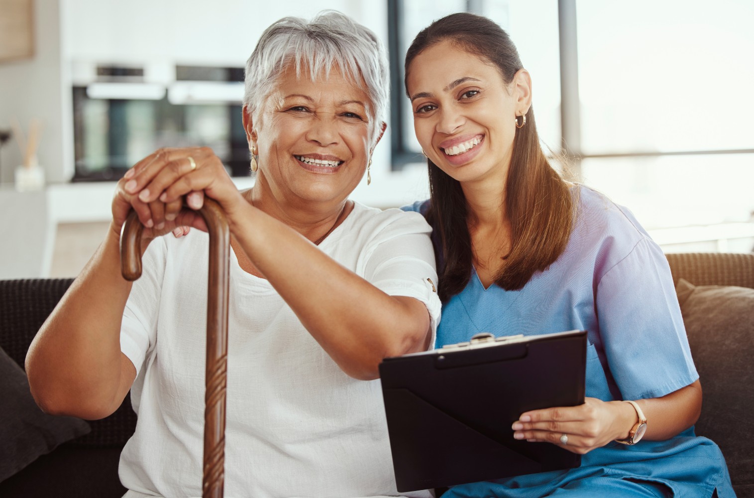 A senior woman with a cane standing beside a nurse holding a clipboard, both smiling