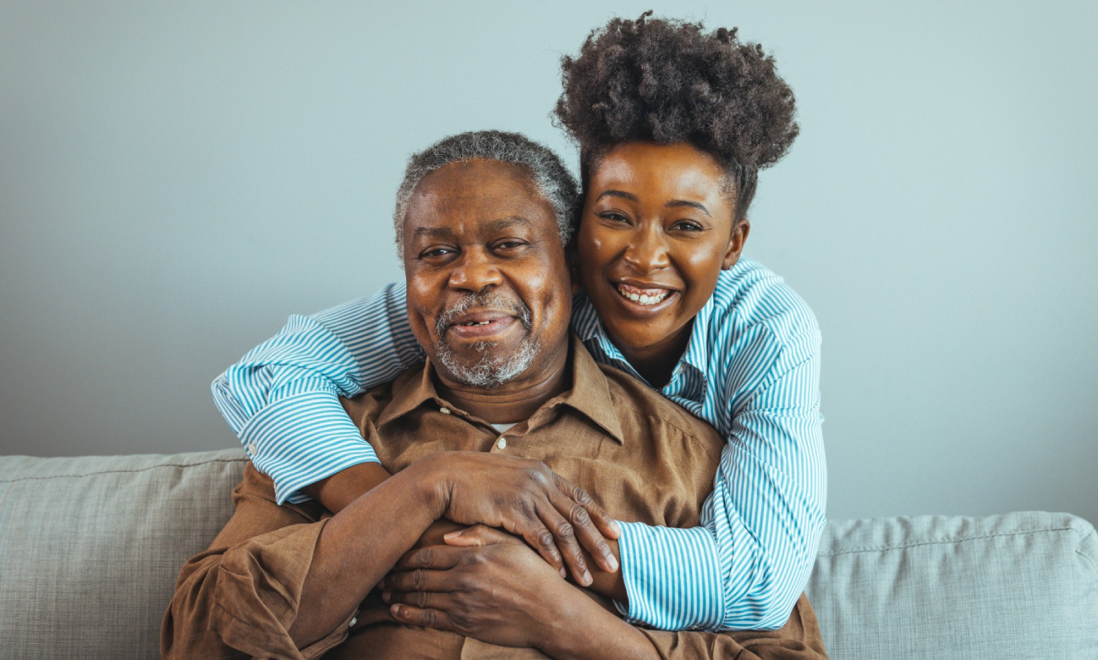 A smiling elderly man and a younger woman hugging on a sofa at home