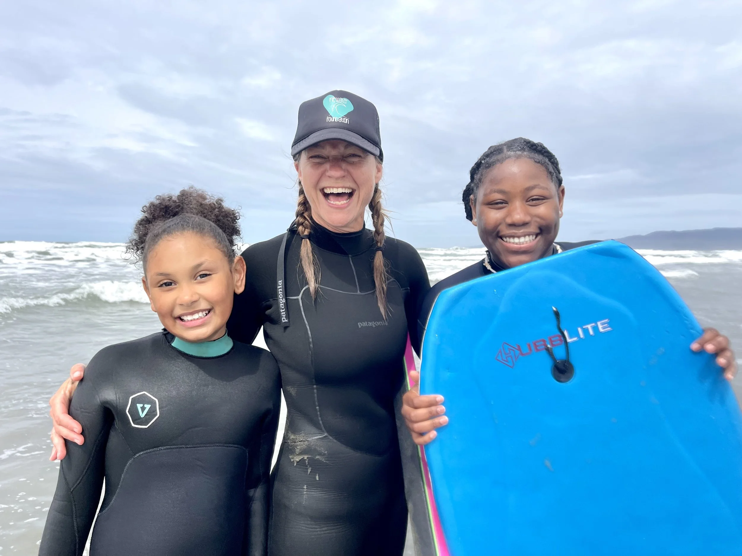 An instructor in a MeWater cap smiling on the beach with two young girls holding a bodyboard