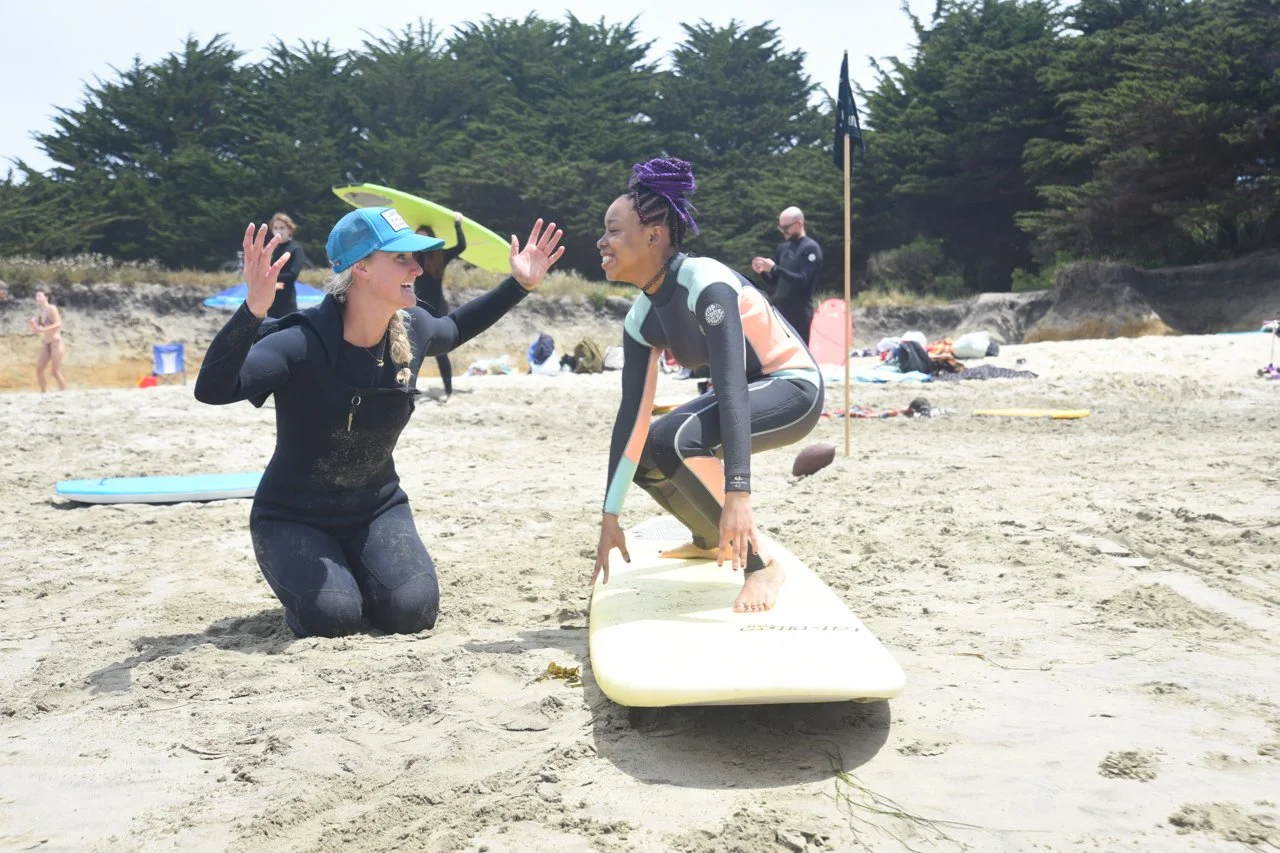 A volunteer instructor high-fiving a young girl practicing her surf stance on the sand