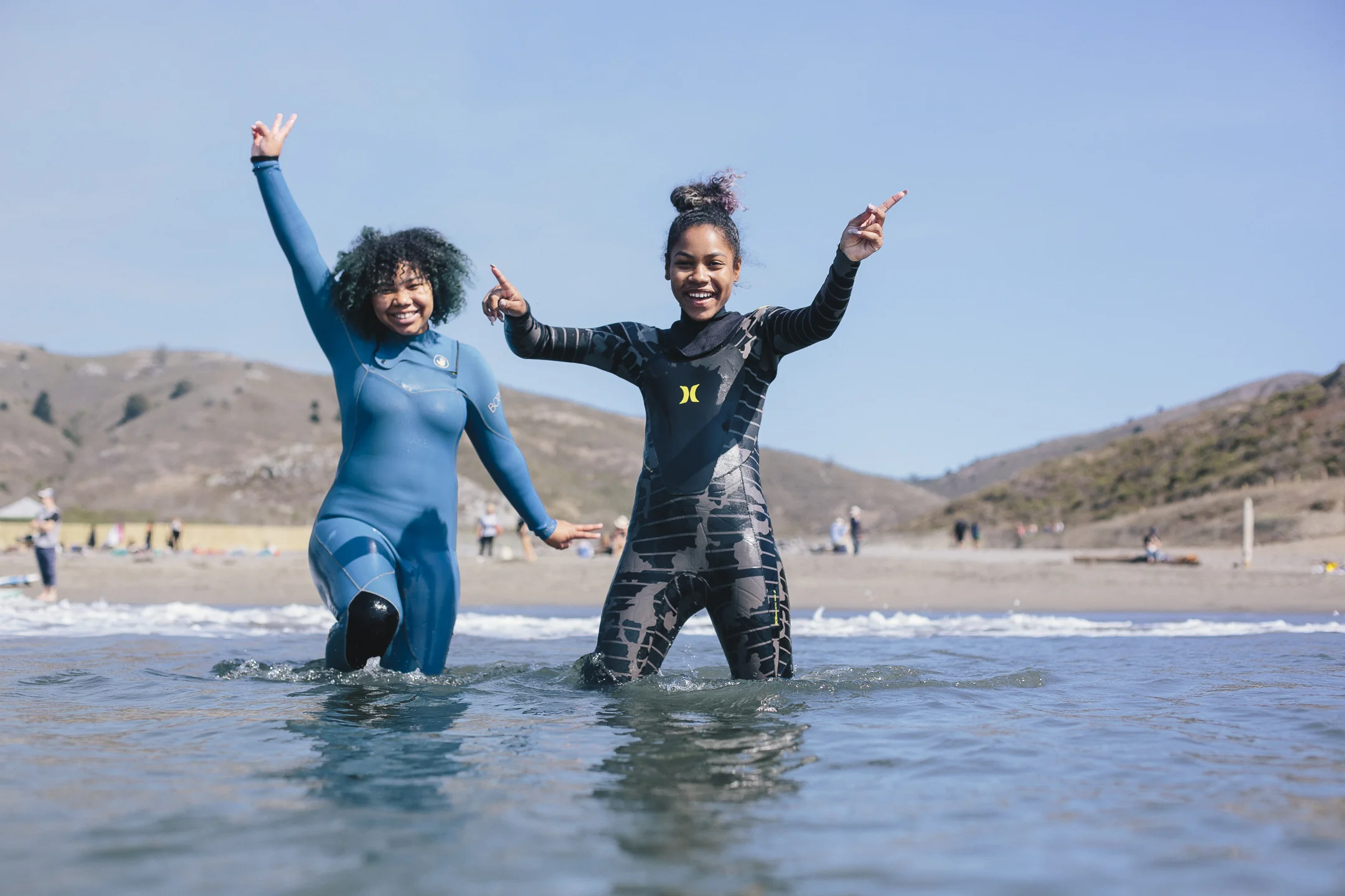 Two teenage girls in wetsuits celebrating with arms raised in shallow ocean water