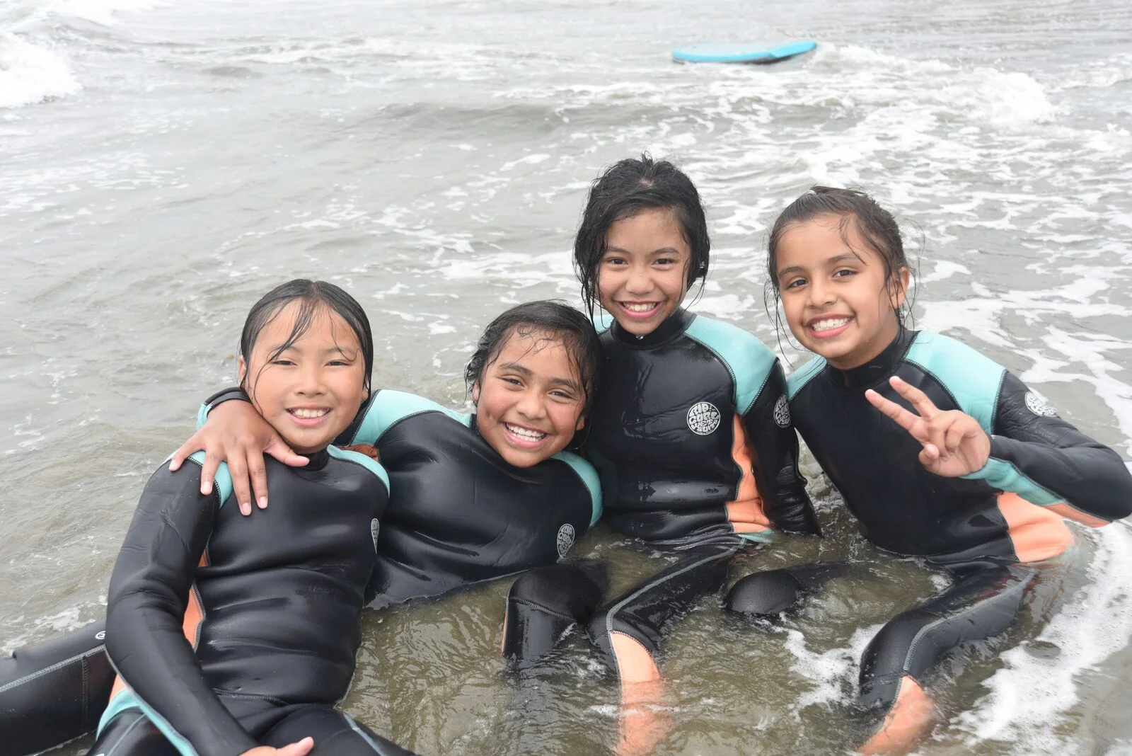 Four young girls in matching wetsuits smiling together in shallow ocean surf