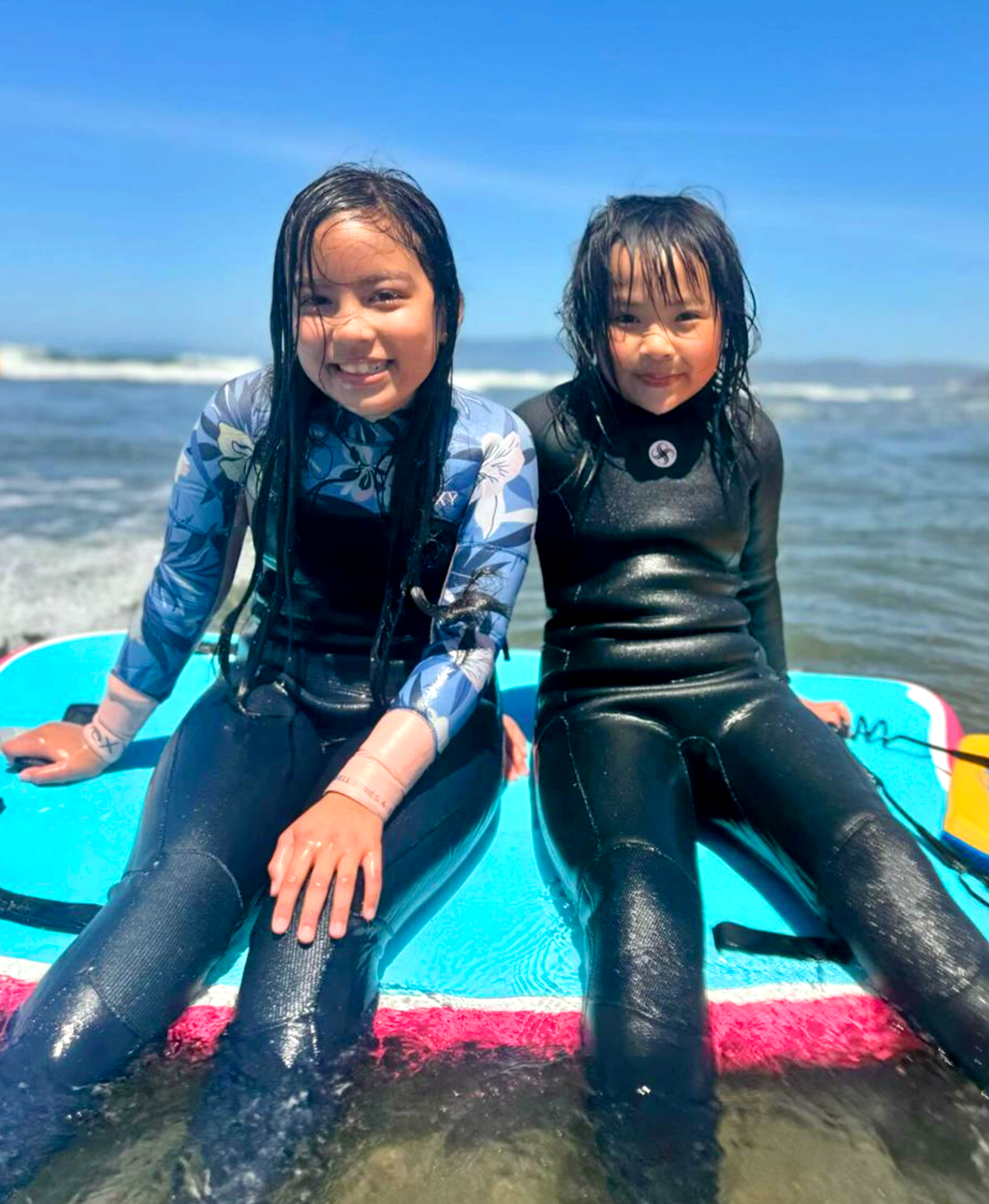 Two young girls in wetsuits sitting together on a surfboard in sunlit ocean water