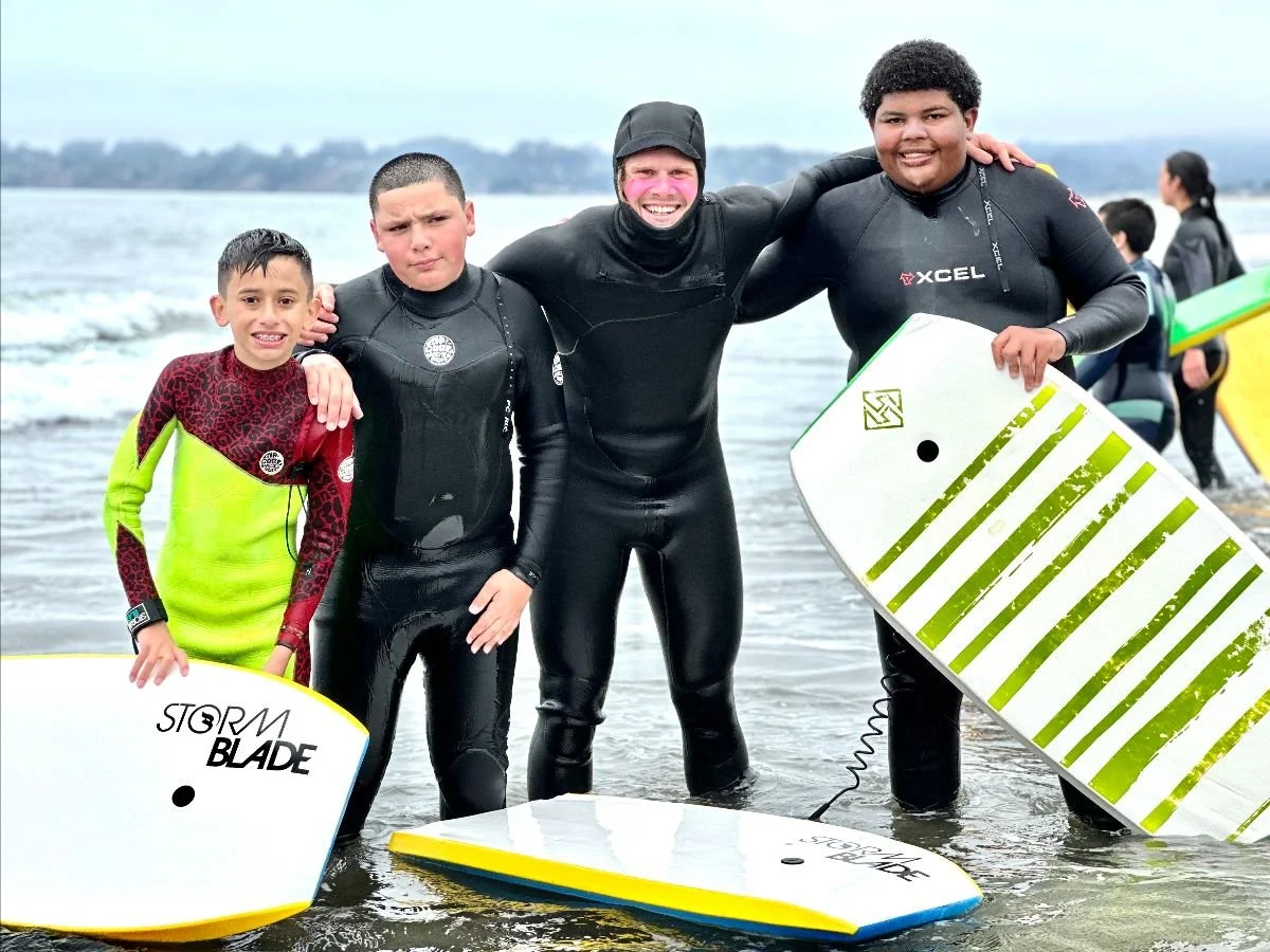 An instructor with his arm around three youth holding bodyboards in the surf