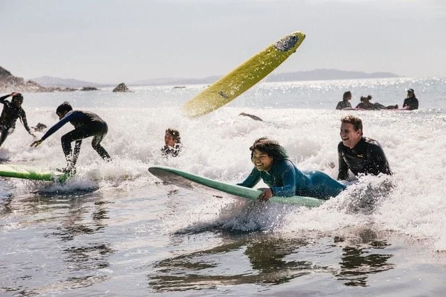 A girl in a blue wetsuit riding a green board prone with an instructor behind her