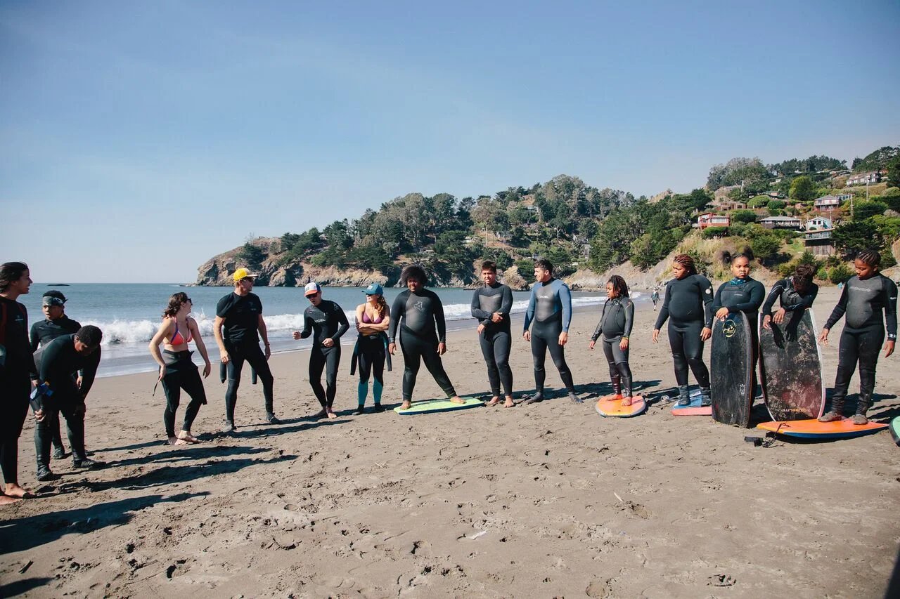 Camp participants standing in a line on the beach with surfboards during an instructor briefing