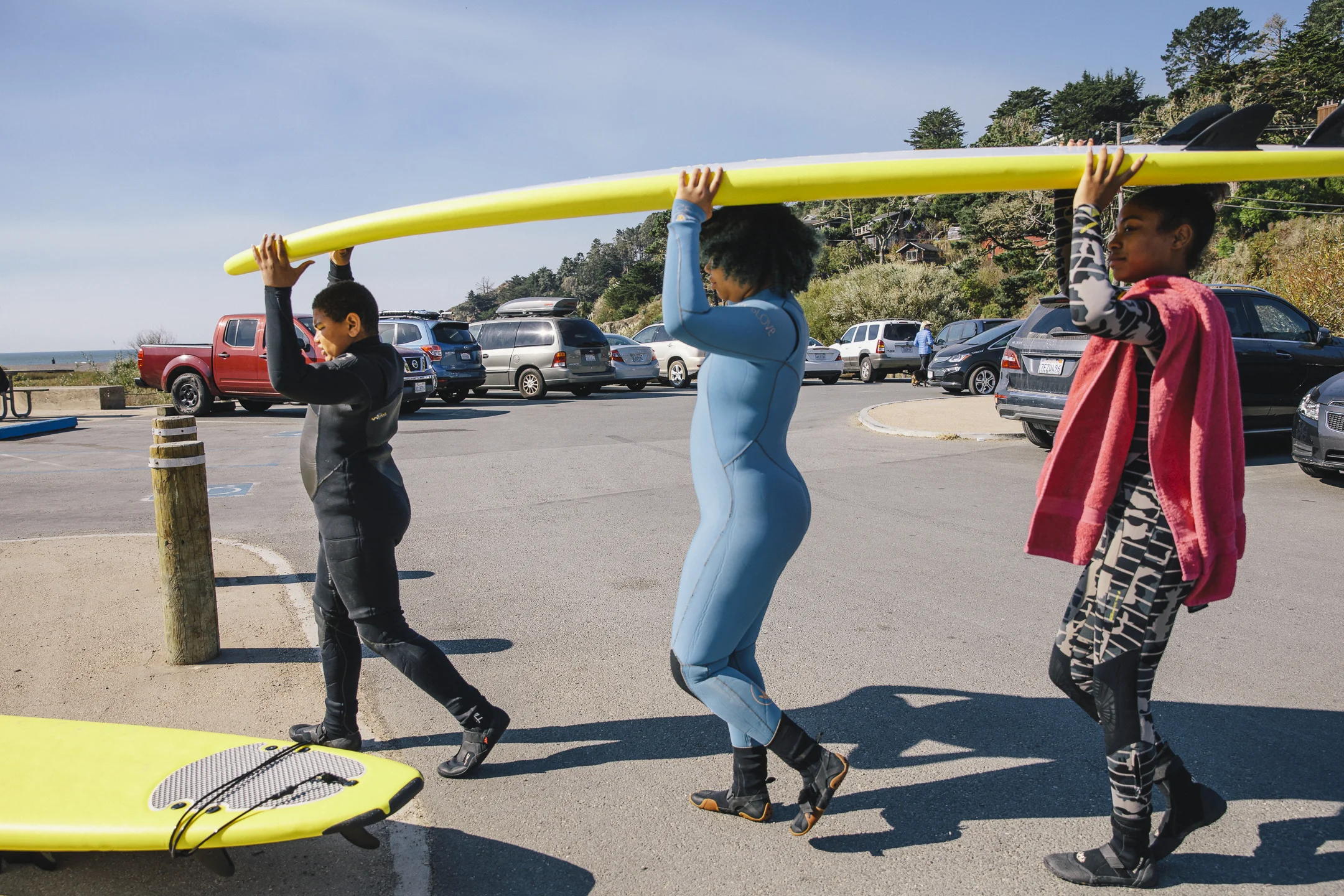 Three youth in wetsuits carrying a yellow surfboard overhead through a beach parking lot